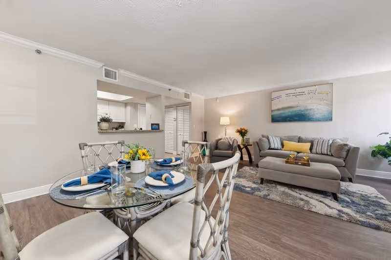A bright and modern living and dining area featuring a round glass dining table set with blue napkins and white plates, four cushioned chairs, a gray sofa with striped and yellow pillows, a matching ottoman, a gray armchair, a floor lamp, a side table with flowers, and a large beach-themed artwork on the wall. The kitchen is visible in the background through a pass-through window.