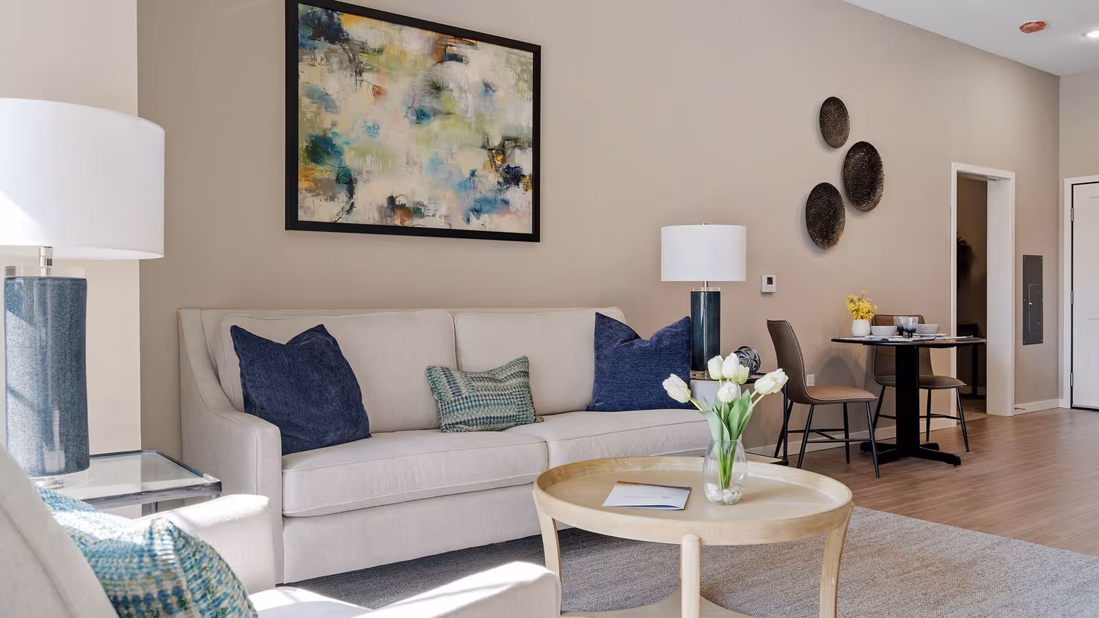 Living room with a beige sofa topped with blue and green pillows, a round coffee table holding a vase of white tulips, wall art, and a small dining table in the background.