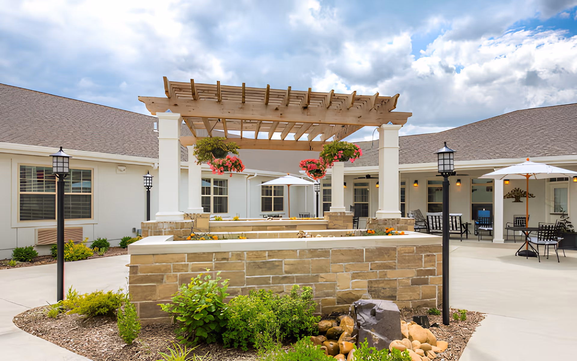 Outdoor courtyard with a wooden pergola, hanging flower baskets, stone planter walls, and patio seating with umbrellas.