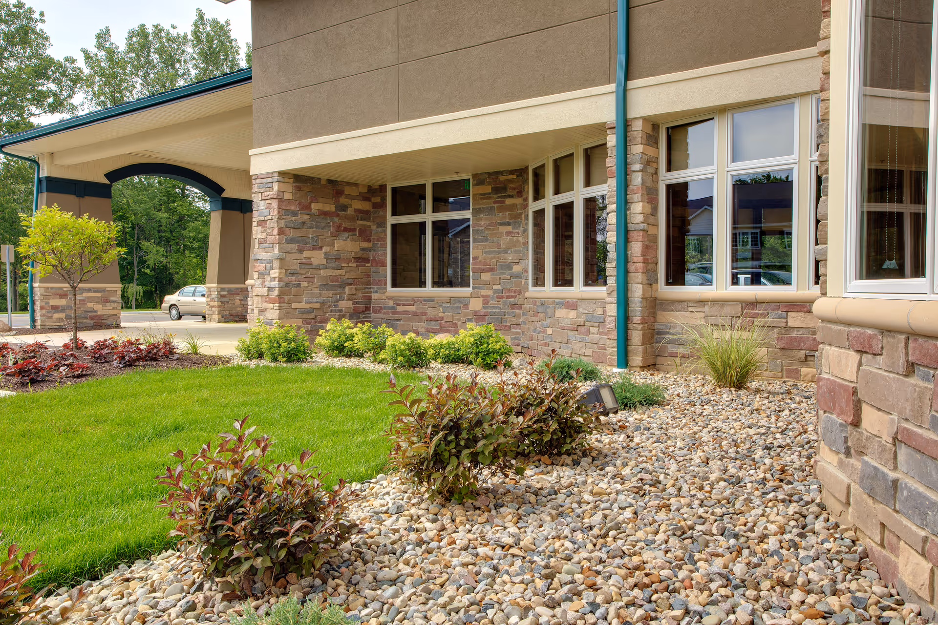 Front exterior of a building with stone veneer, large windows, a covered entrance, and landscaped lawn with decorative rock beds.