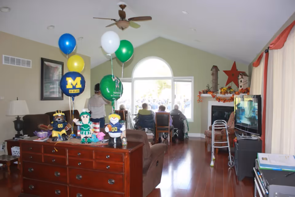 A senior living facility common area with several elderly residents seated facing a large window. The room has wooden floors, a ceiling fan, a TV, and a dresser with balloons and stuffed sports mascots on top. The window lets in natural light, and there are fall-themed decorations on the fireplace mantel.