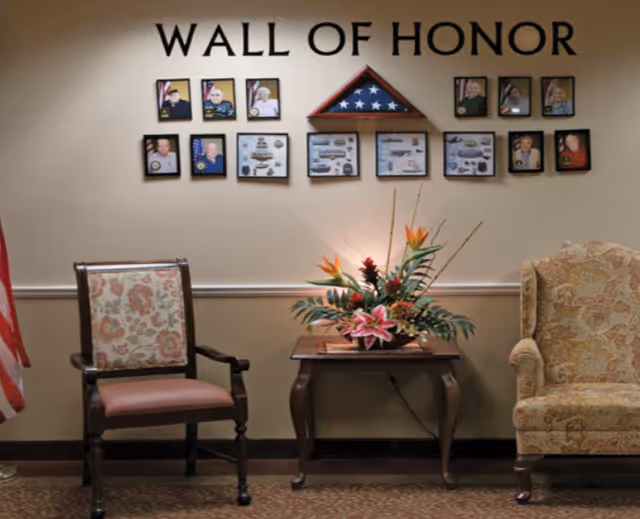Seating area with two chairs and a small table topped with a floral arrangement beneath a 'Wall of Honor' display of framed photos and a folded American flag.