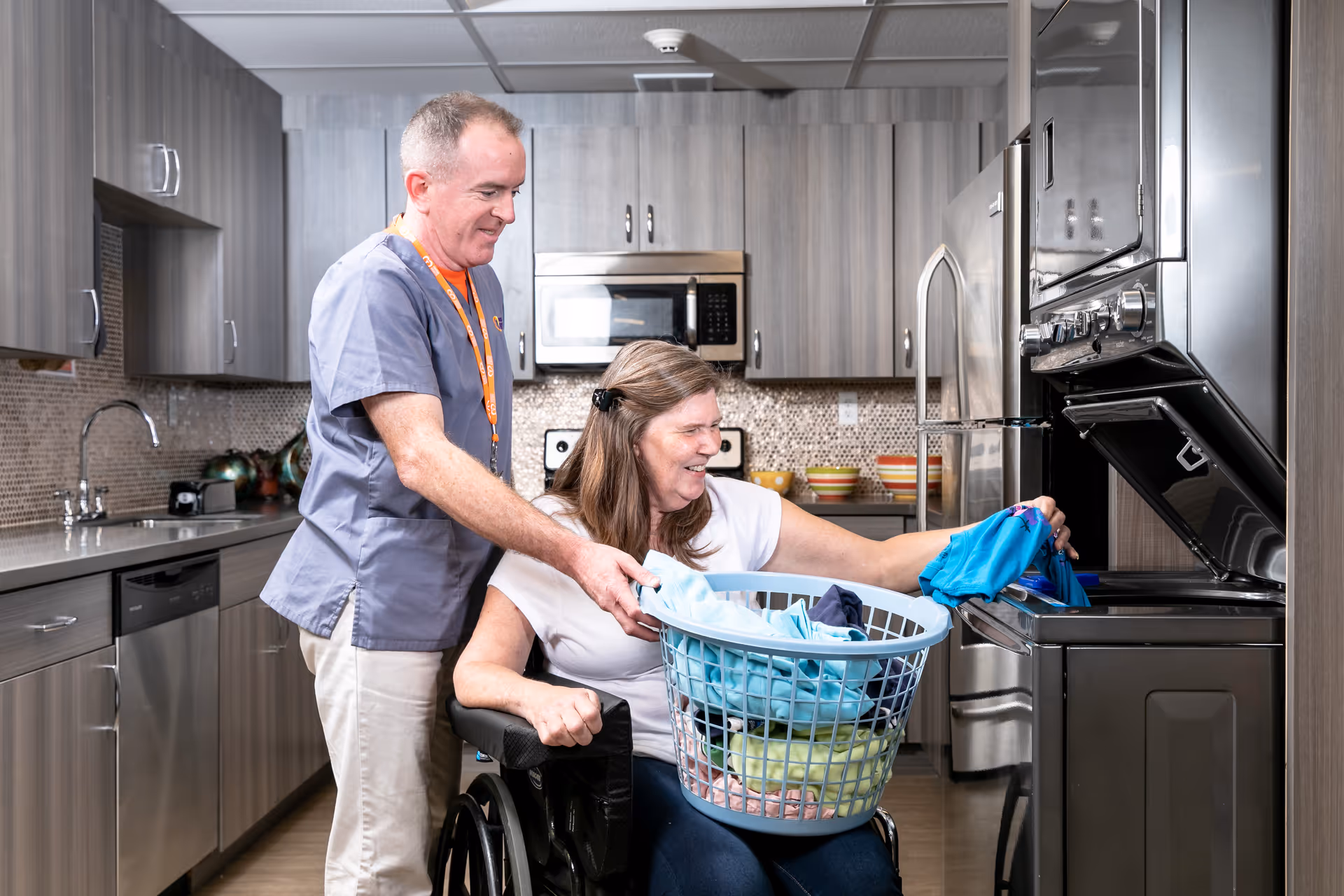 A man in scrubs assists a woman in a wheelchair as she loads clothes into a front-loading washing machine in a modern kitchen with wooden cabinets and stainless steel appliances.
