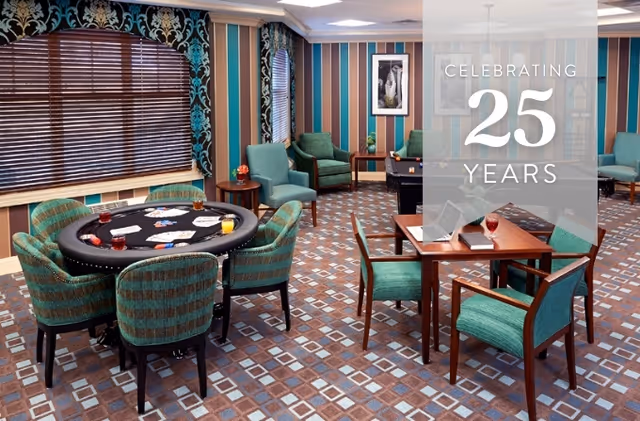 A cozy game room with a poker table surrounded by green upholstered chairs, a smaller table with two chairs, and additional seating along the walls. The room features patterned carpet, striped wallpaper, and large windows with wooden blinds and decorative curtains.