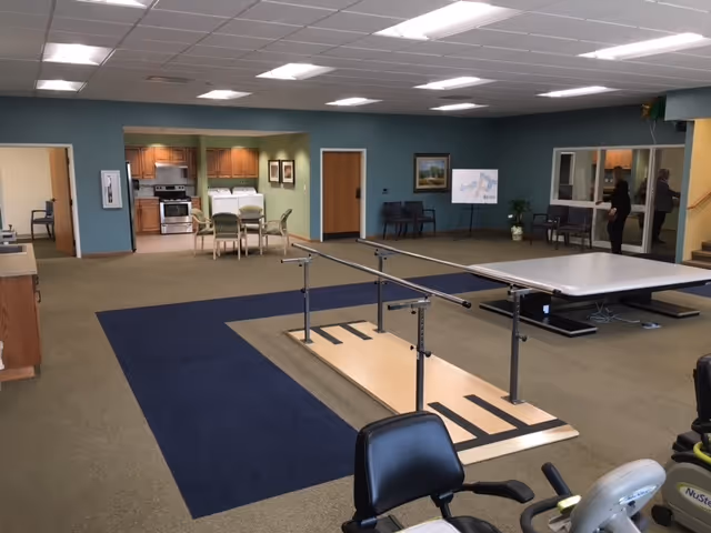 Interior view of a senior living facility room with a kitchen area in the background, a table with chairs, parallel bars for physical therapy or rehabilitation in the center, and exercise equipment in the foreground. The room has a blue and beige carpet, fluorescent ceiling lights, and a few chairs along the walls.