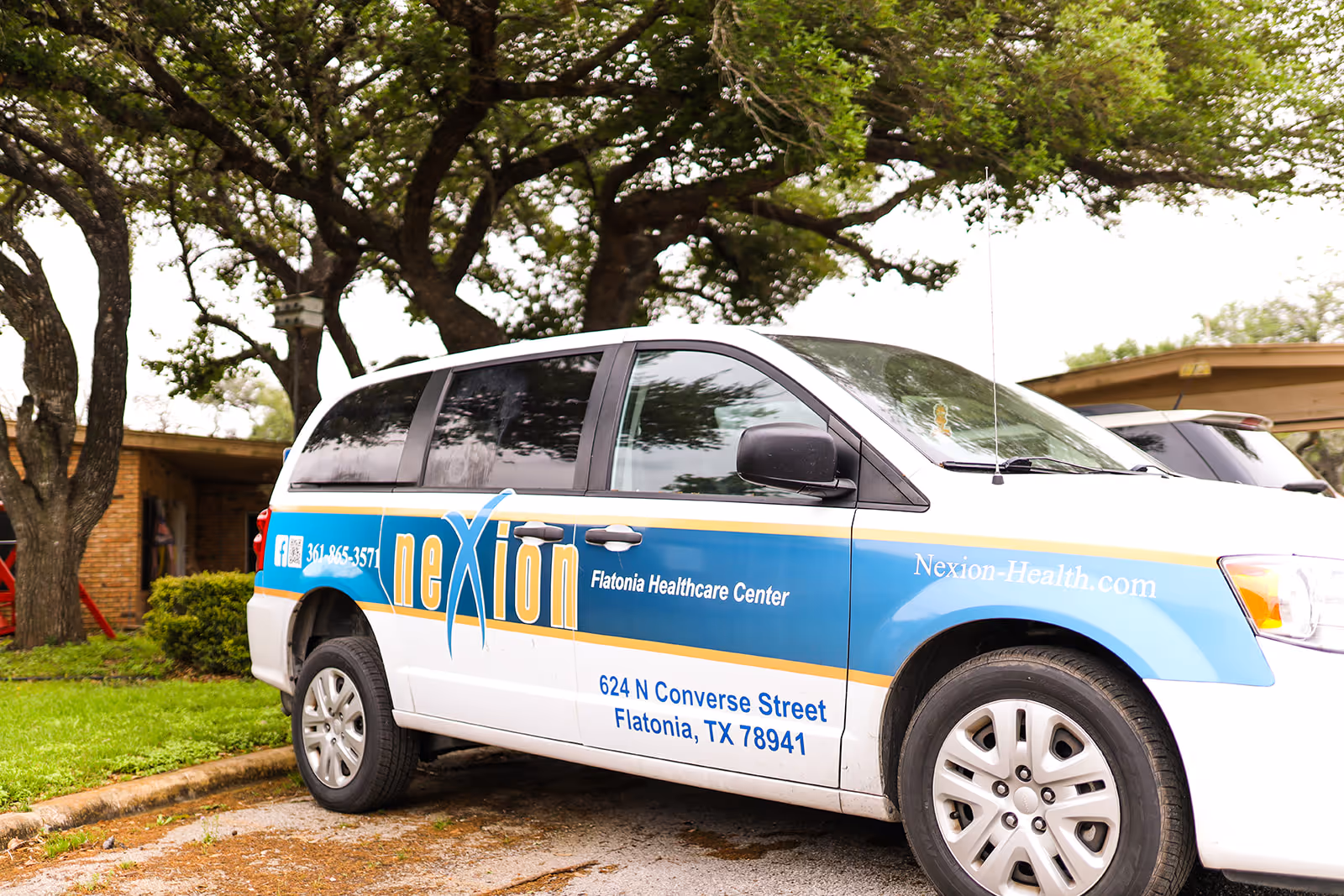 A Nexion-branded minivan parked outside Flatonia Healthcare Center with trees and part of the building visible behind it.