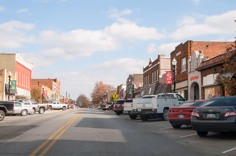 View down a small town street lined with parked cars and brick buildings on both sides under a partly cloudy sky.
