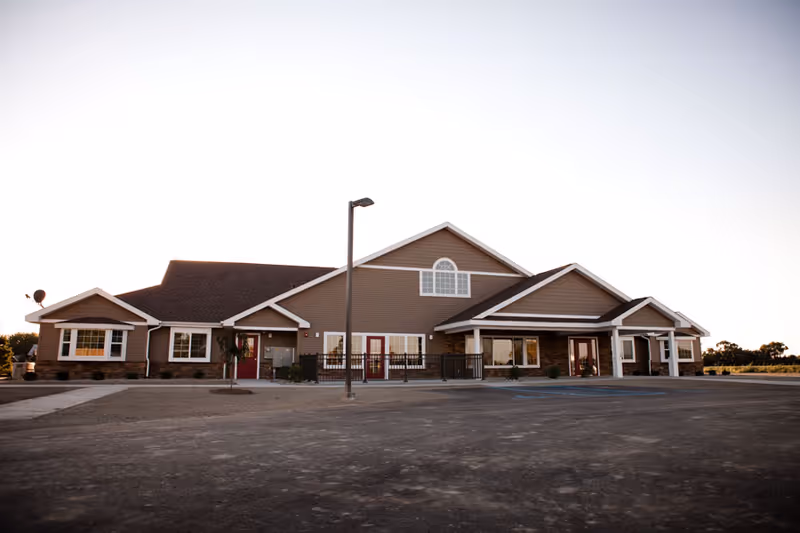 Exterior view of a single-story assisted living facility building with brown siding, white trim, multiple windows, and a covered entrance. The building is surrounded by a paved area and some landscaping with small plants and trees.