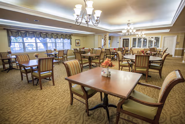 A spacious dining room with multiple wooden tables and cushioned chairs arranged neatly. Each table has a small floral centerpiece. The room features beige walls, carpeted floors with a patterned design, large windows with valances, and elegant chandeliers hanging from the ceiling providing warm lighting.
