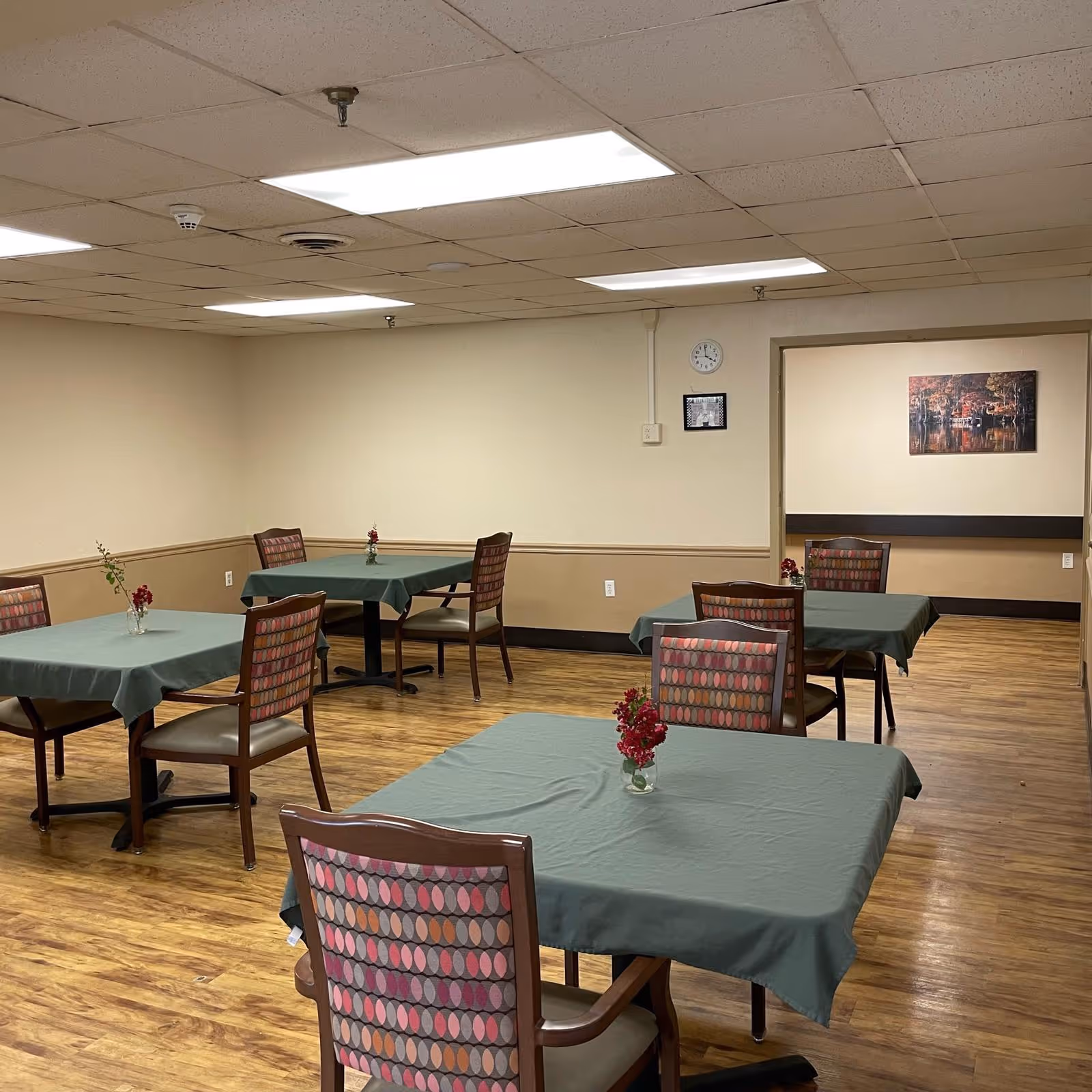 A dining room with several tables covered in green tablecloths, each with a small vase of red flowers. The room has wooden flooring, beige walls with a chair rail, and a drop ceiling with fluorescent lighting. There is a clock and a framed picture on the far wall, and another room with similar decor is visible through an open doorway.