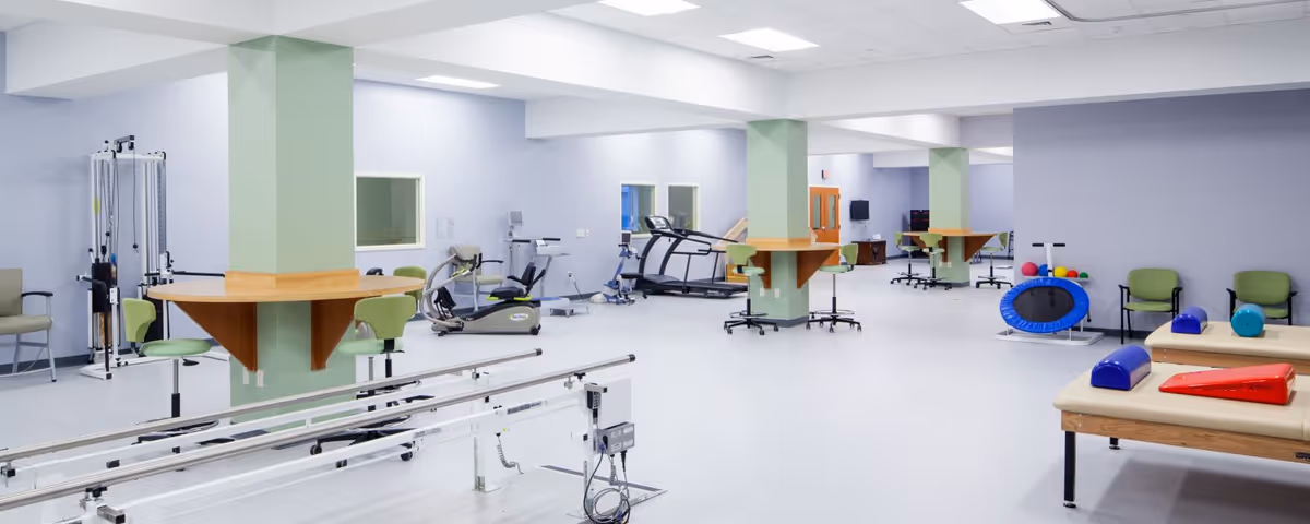 A spacious rehabilitation therapy room with exercise equipment including parallel bars, stationary bikes, treadmills, therapy tables with cushions, chairs, and therapy balls. The room has light-colored walls and floors, green support columns with wooden ledges, and bright overhead lighting.