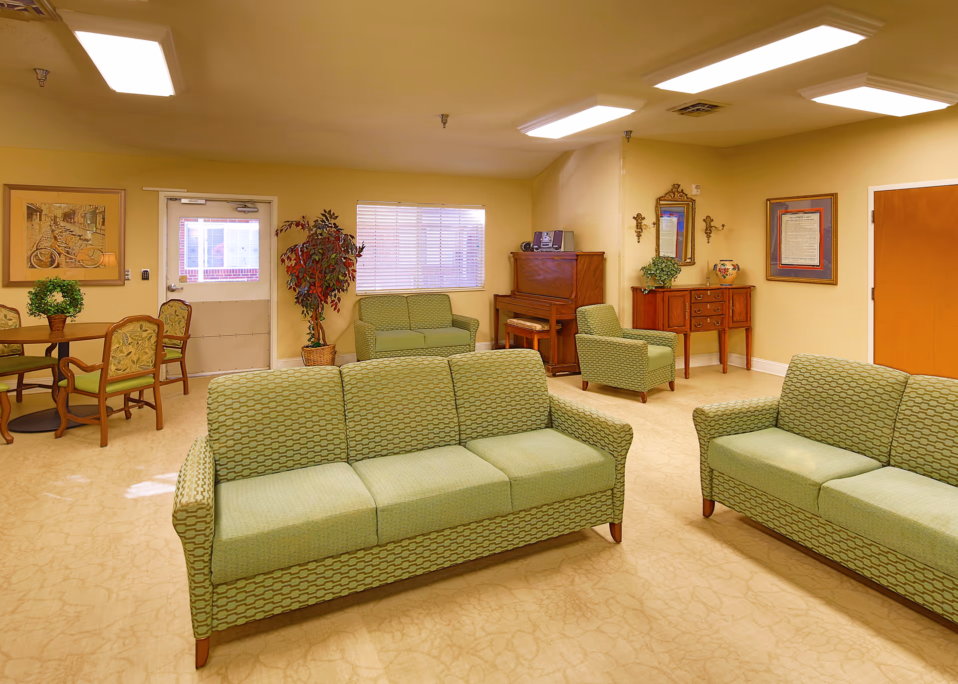 A bright common room with green patterned sofas and chairs, a piano, and a small dining table.