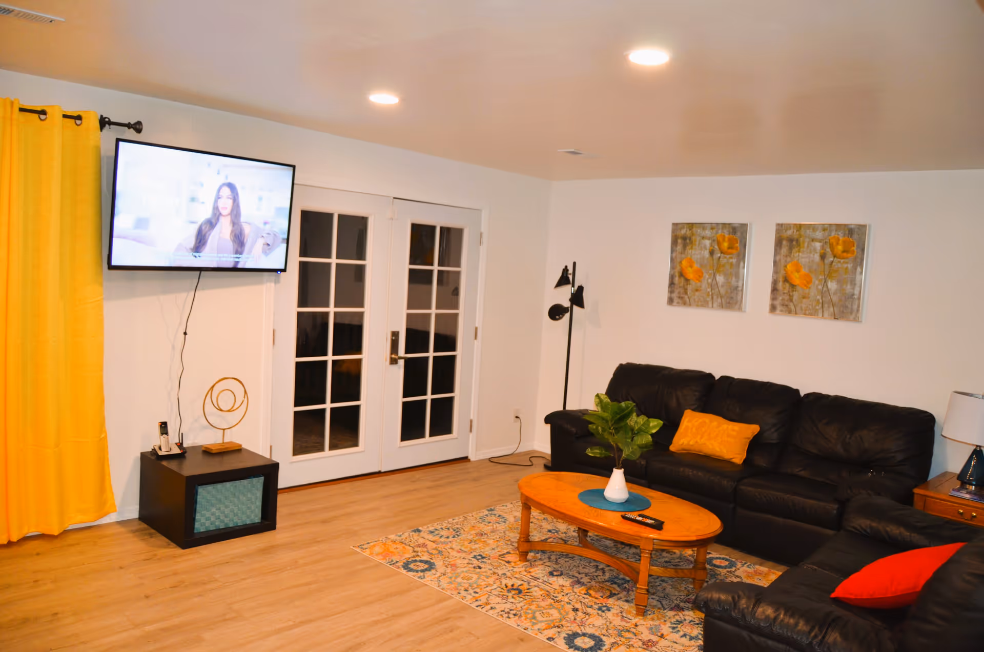 Cozy living room with black leather sofas, a wooden coffee table, wall-mounted TV, yellow curtains, and French doors.