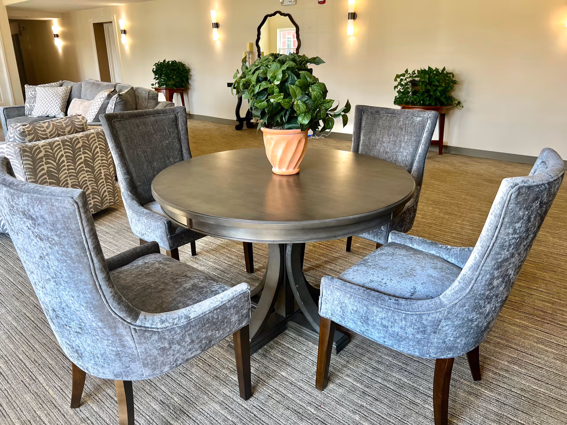 Round wooden table with a potted plant surrounded by four upholstered chairs in a lounge area with sofas and plants in the background.