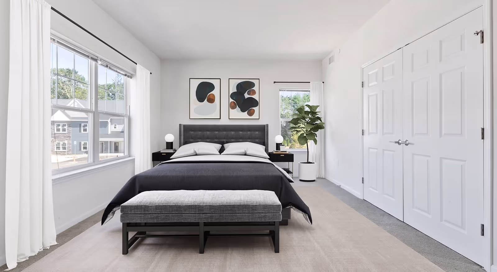 A modern bedroom with a large bed featuring a dark gray upholstered headboard and black bedding. Two abstract art pieces hang above the bed. There are two black nightstands with round white lamps on each side of the bed. A gray bench is positioned at the foot of the bed. The room has large windows with white curtains, a potted plant near one window, and white double closet doors on the right side.