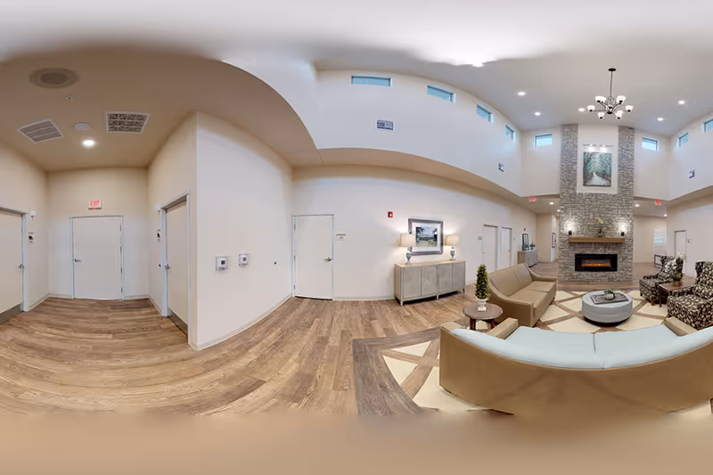 A spacious and well-lit common area in a senior living facility featuring a high ceiling with small windows near the top. The room has a stone fireplace with a painting above it, surrounded by wall sconces. There are several seating options including a light blue sofa, a brown sofa, and patterned armchairs arranged around a round coffee table. The floor has a wood and tile design, and there is a sideboard with two lamps and a framed picture on the wall. Multiple doors lead to other rooms, and the walls are painted a light beige color.