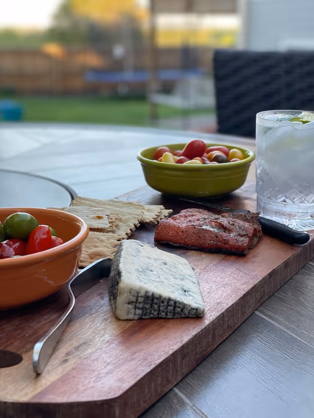 A wooden serving board on a table with a wedge of blue cheese, a piece of cured meat, crackers, a bowl of mixed olives and cherry tomatoes, and a glass of ice water with a lime wedge. The background shows an outdoor setting with blurred greenery and patio furniture.