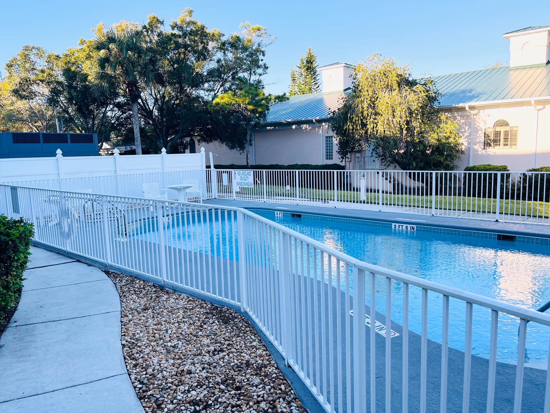 Fenced outdoor swimming pool with chairs, a pathway and landscaping in front of a light-colored building and trees.