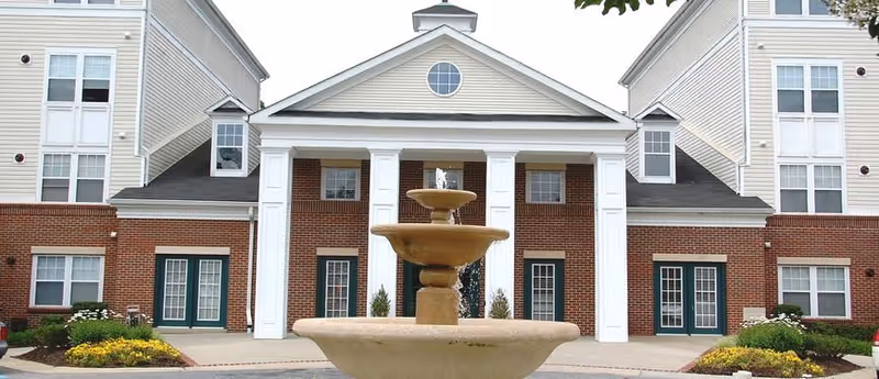 Front exterior view of a senior living facility building with a central entrance featuring white columns and a triangular pediment. In front of the entrance is a large tiered water fountain surrounded by landscaped flower beds and shrubs.
