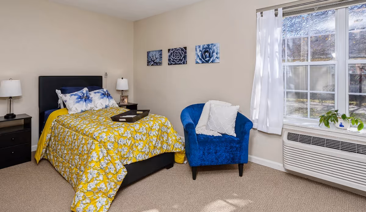 A bright bedroom with a bed dressed in yellow floral bedding, a blue armchair, nightstands, and a large window with an air conditioning unit.