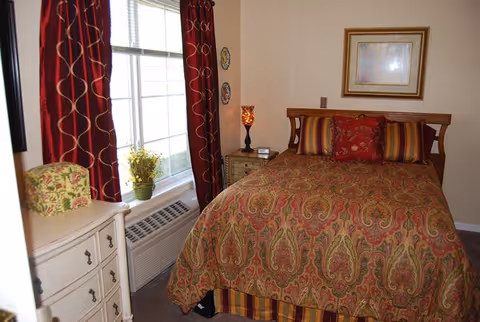 A cozy bedroom with a double bed covered in a patterned red and gold bedspread, matching pillows, and a wooden headboard. To the right of the bed is a small nightstand with a decorative lamp and a clock. A framed picture hangs on the wall above the bed. On the left side of the room, there is a window with red curtains featuring a wavy gold pattern, a white dresser with a floral cushion on top, and a potted plant on the windowsill. Below the window is an air conditioning unit.