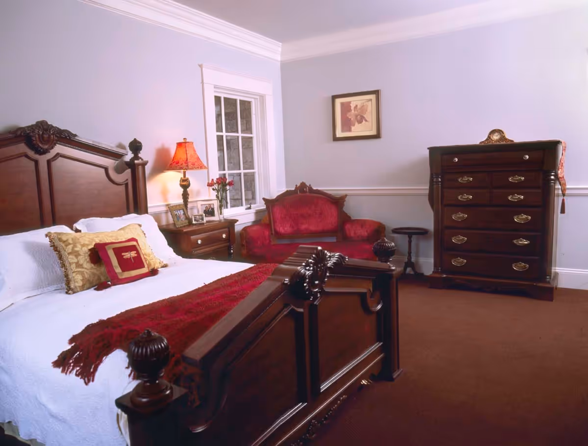 A bedroom with classic wooden furniture including a large bed with white bedding and decorative pillows, a nightstand with a lamp and framed photos, a red upholstered loveseat, a small round side table, and a tall dresser with brass handles. The walls are light-colored with a framed picture hanging above the loveseat.