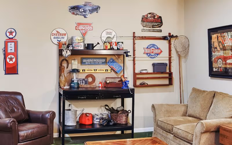 A cozy living room corner featuring a brown leather armchair and a beige fabric sofa. Between them is a black workbench with various tools and vintage signs on the wall above it, including gas station logos and a sign that reads 'Grandpa's Workshop'. A fishing net and a framed picture are also visible on the walls.