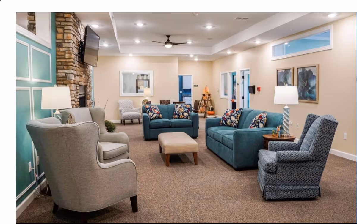 Spacious furnished common lounge with teal sofas, patterned armchairs, an ottoman, lamps and a stone fireplace.