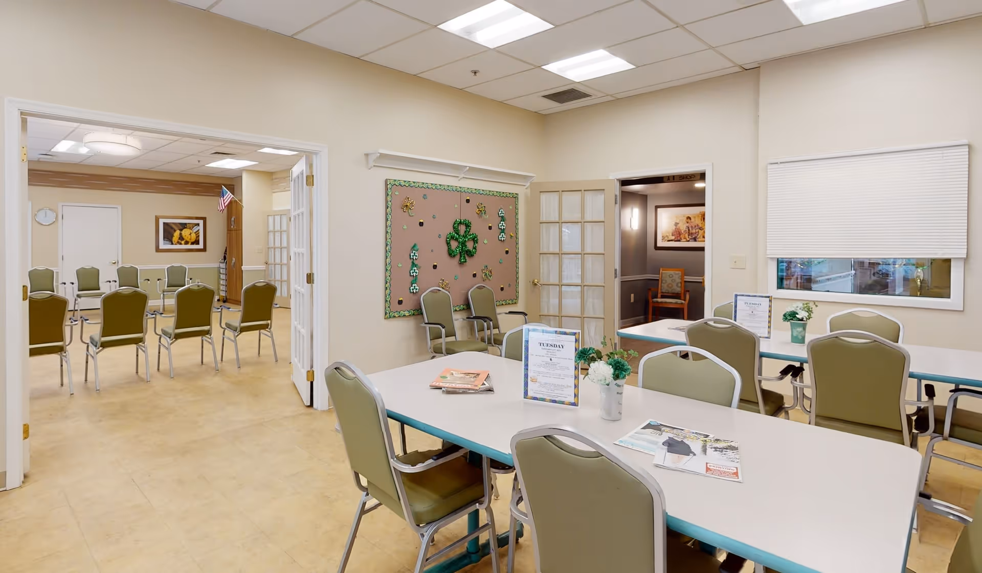 Interior view of a community room in Arden Courts - ProMedica Memory Care Community. The room has several tables with green cushioned chairs arranged around them. There are decorations on the walls including a bulletin board with shamrock decorations. Through an open double door, another room with more chairs arranged in rows is visible. The room is well-lit with ceiling lights and has a window with a closed blind.