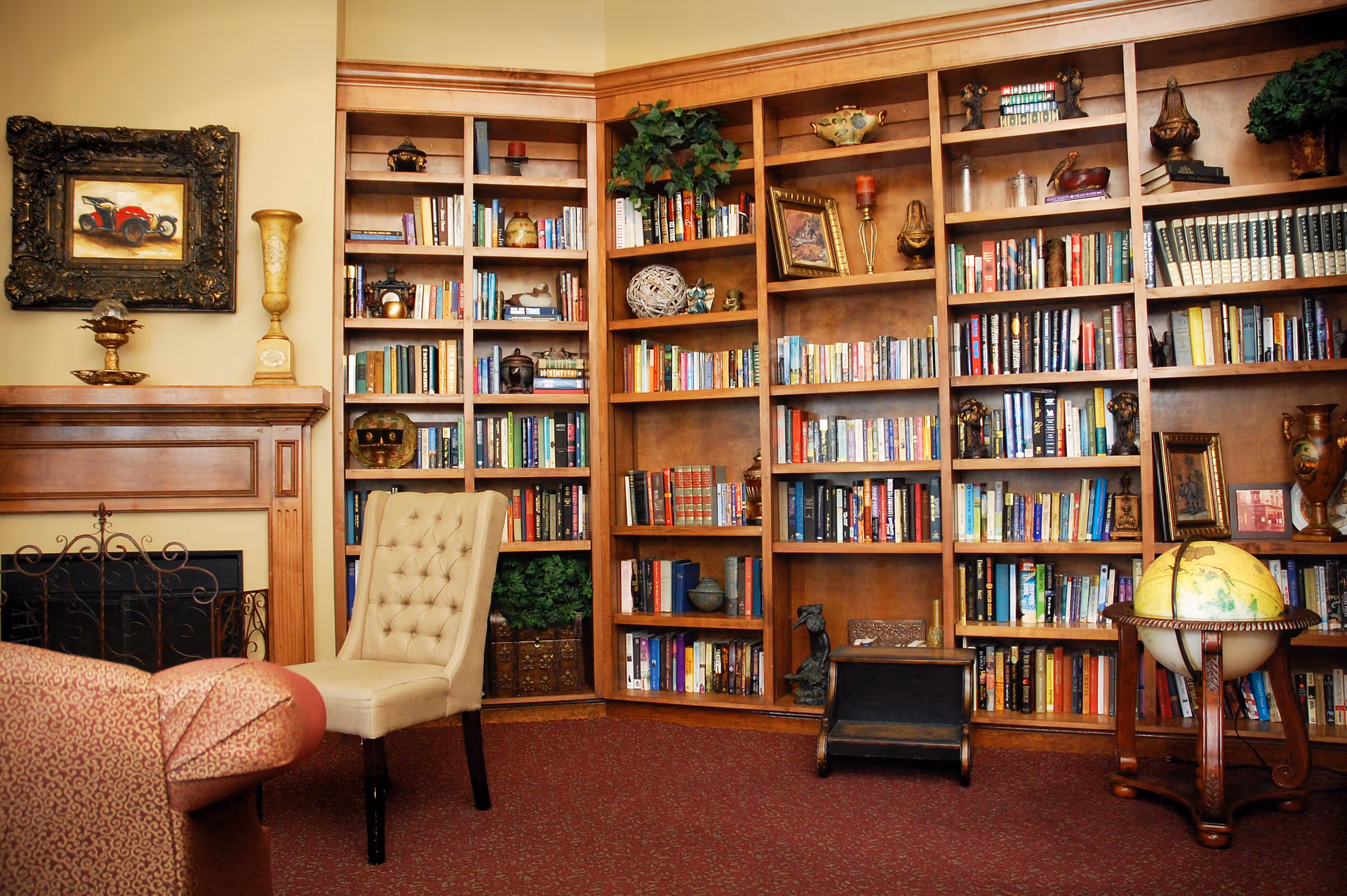 A cozy library room with wooden bookshelves filled with books and decorative items, a beige tufted chair, a globe on a wooden stand, and a fireplace with a decorative screen and framed artwork above it.