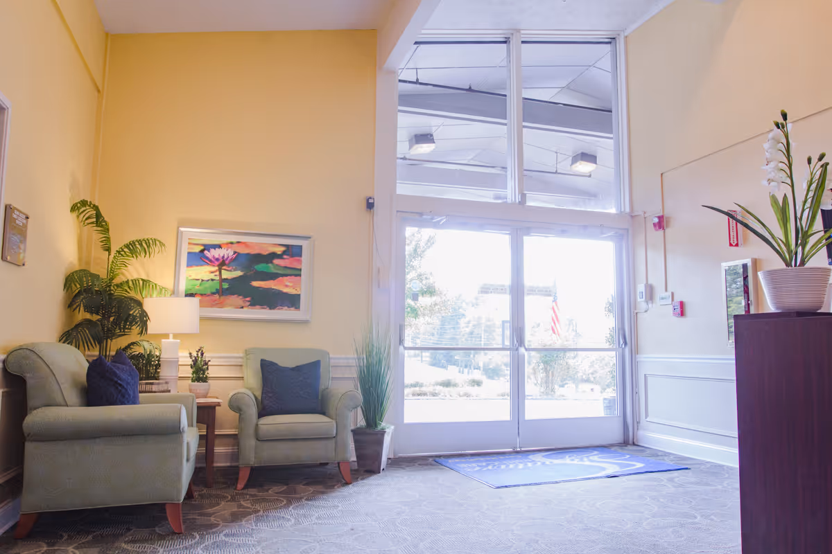 A bright and welcoming lobby area with two green armchairs, a small wooden side table with a lamp, and potted plants. There is a colorful painting on the yellow wall and large glass double doors letting in natural light.