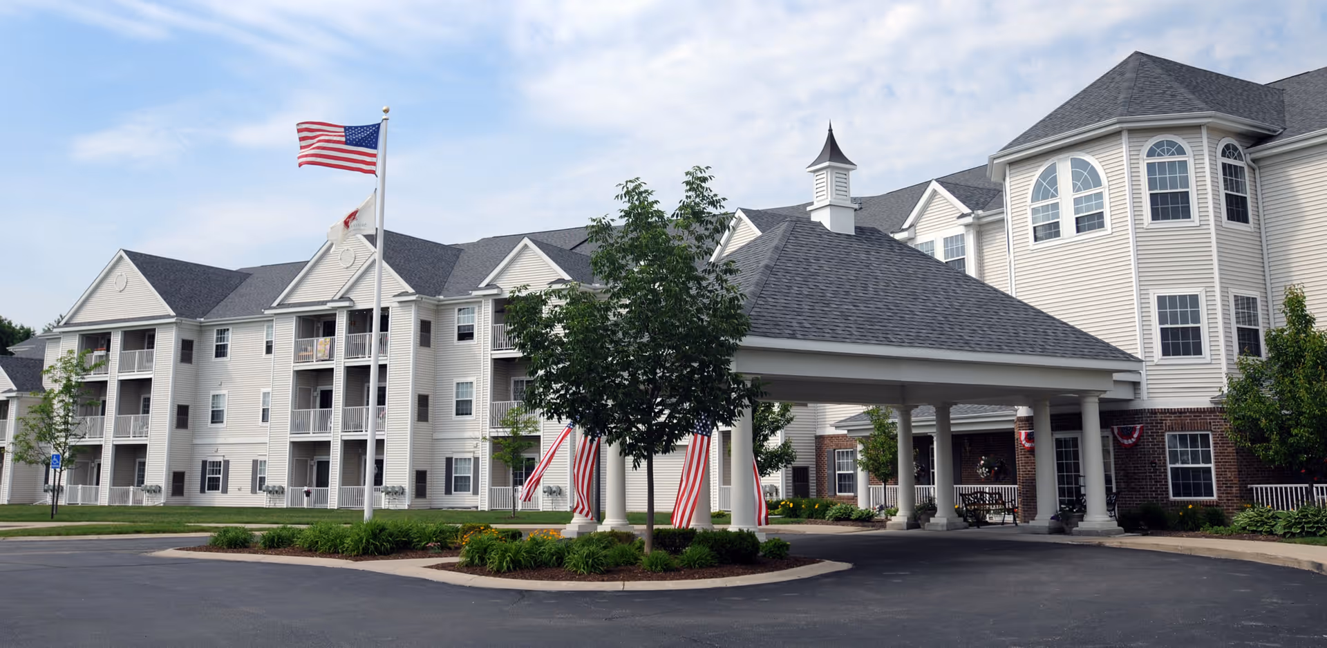 Exterior view of a large, multi-story retirement community building with beige siding and brick accents. The building features multiple windows, balconies, and a covered entrance supported by white columns. There are American flags displayed near the entrance and a flagpole with the American flag and another flag. The surrounding area includes a paved driveway, green lawns, and some trees.