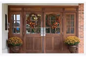 Double wooden entrance doors with glass panels decorated with autumn wreaths. Two large pots of yellow chrysanthemums flank the doorway, and the exterior wall is made of red brick.