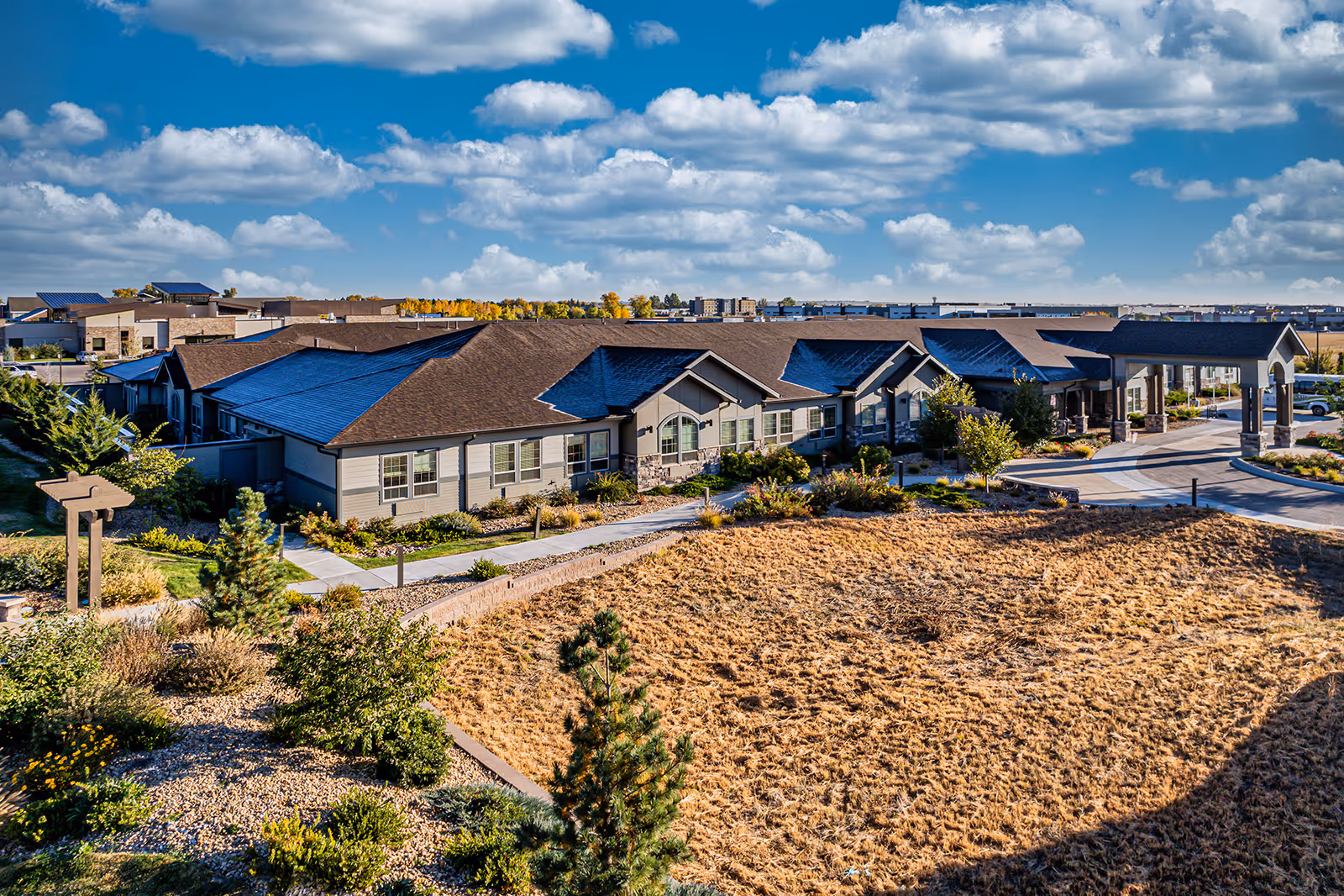 Aerial view of a single-story senior living facility building with a brown roof and beige siding, surrounded by landscaped gardens and dry grass under a partly cloudy blue sky.