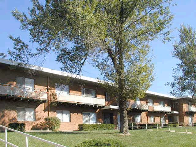 Exterior view of a two-story brick apartment building with balconies and windows, surrounded by green grass and trees under a clear blue sky.