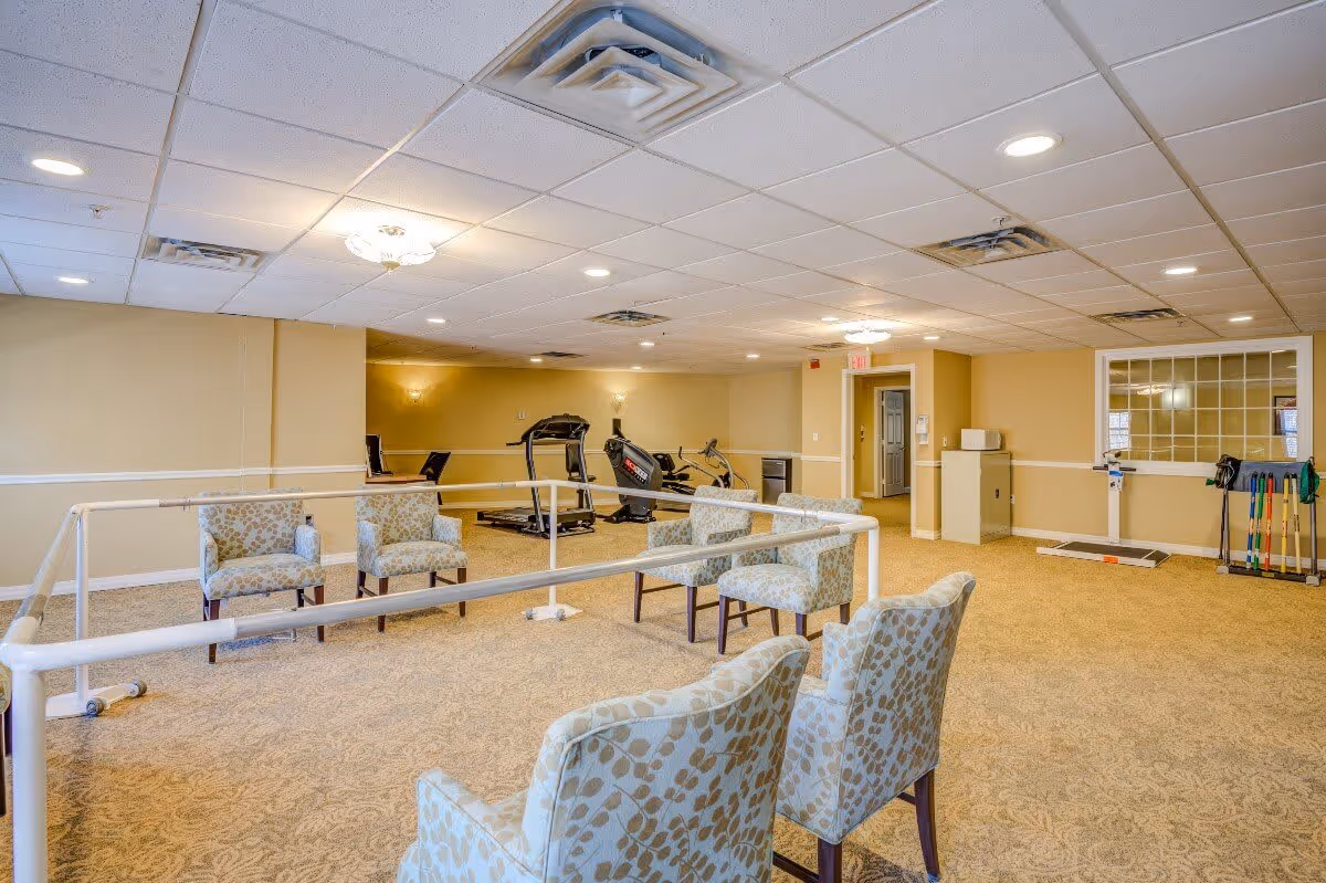 A spacious room with beige walls and carpeted floor featuring several patterned armchairs arranged around a white handrail structure. In the background, there are exercise machines including a treadmill and a stationary bike. The ceiling has recessed lighting and air vents, and there is a window with multiple panes on the right wall.