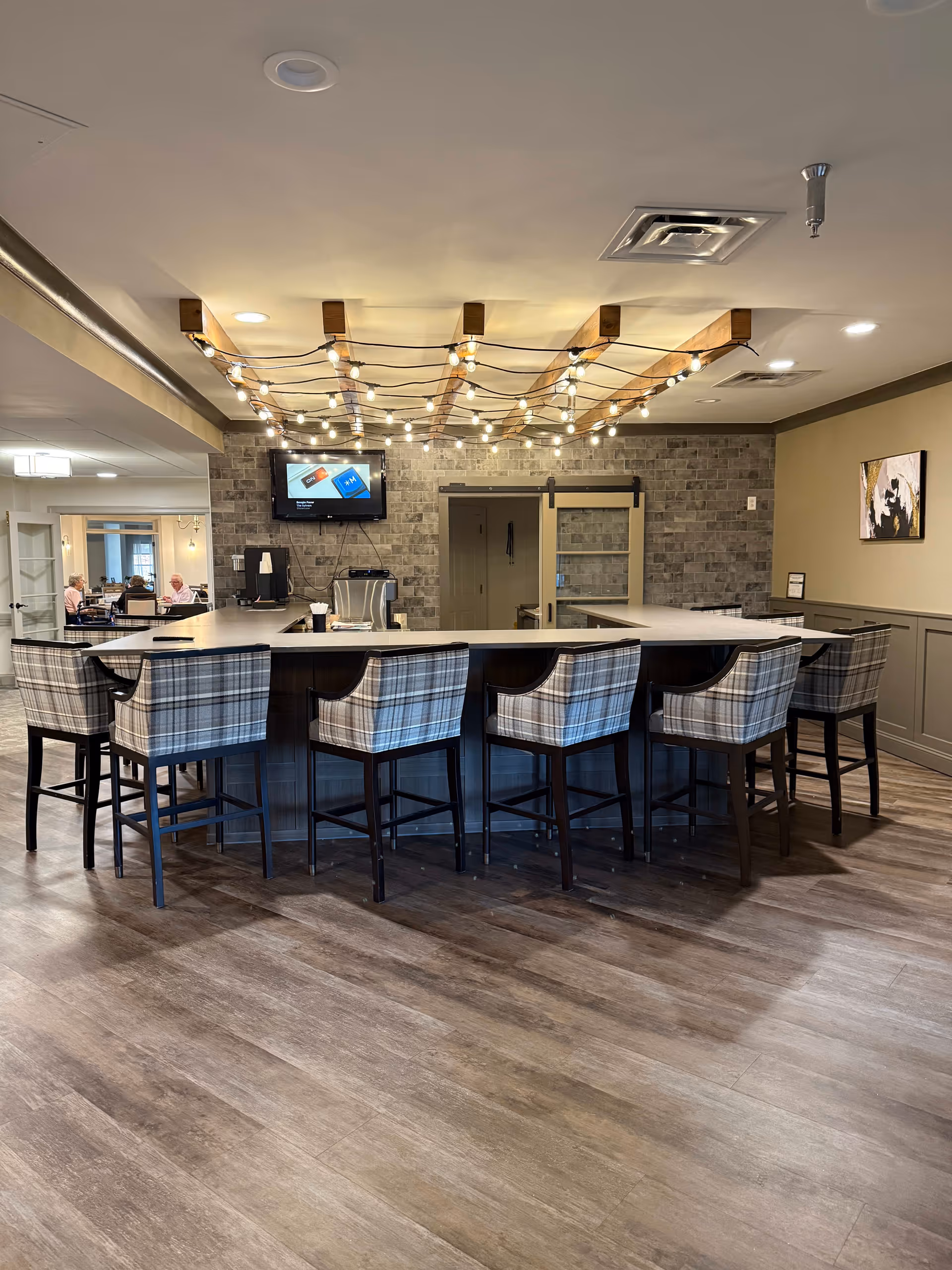 Communal bar-style dining area with plaid high-back stools around a central island under hanging string lights.