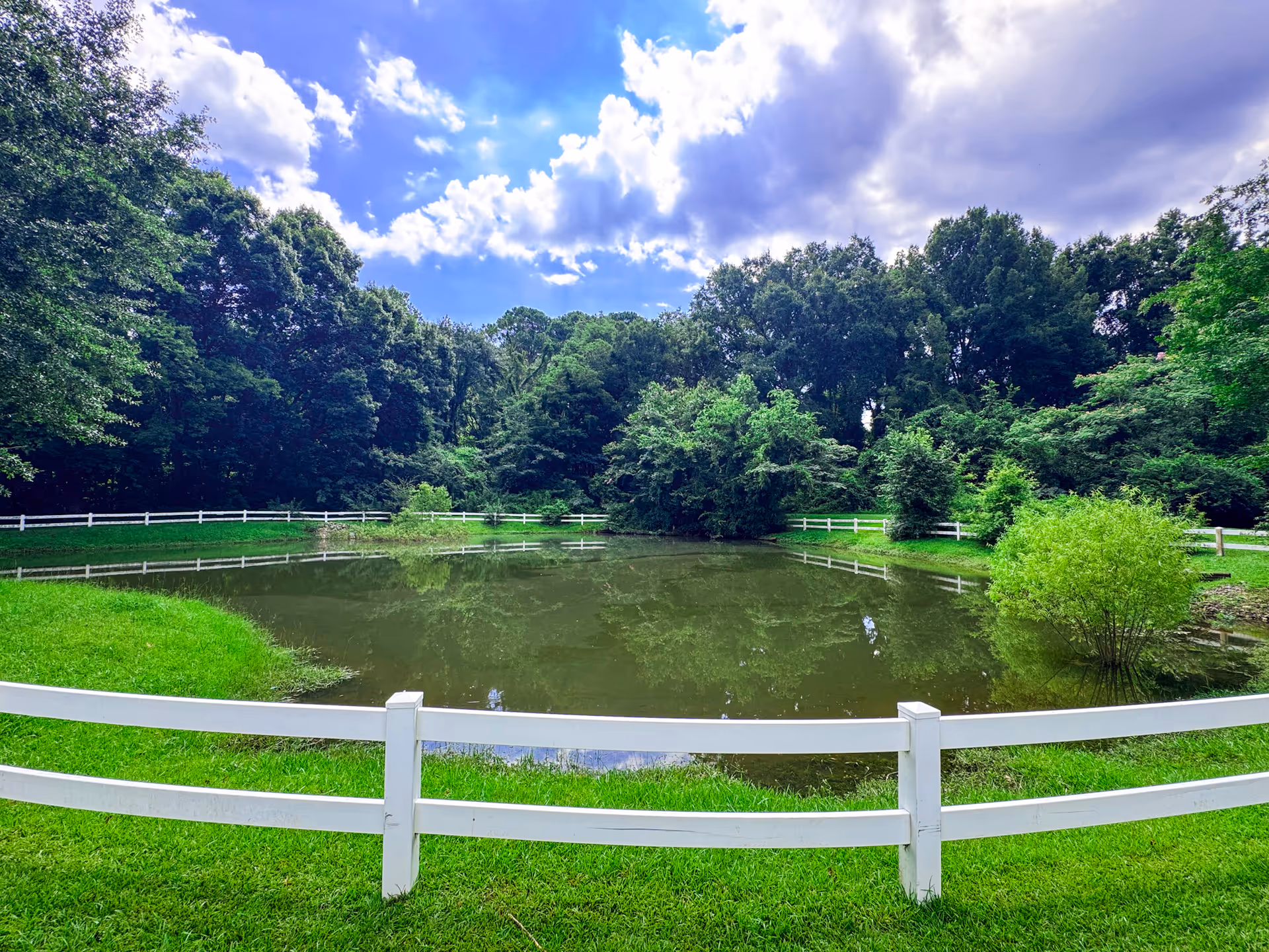 A small pond surrounded by a white wooden fence with lush green grass and dense trees in the background under a partly cloudy blue sky.