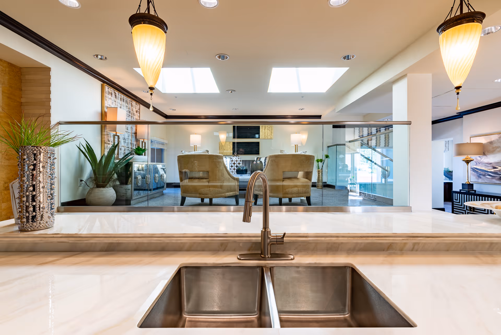 View from a kitchen sink area with a modern stainless steel faucet and double basin sink, looking out into a bright living room with beige armchairs, glass railing, plants, and ceiling skylights.