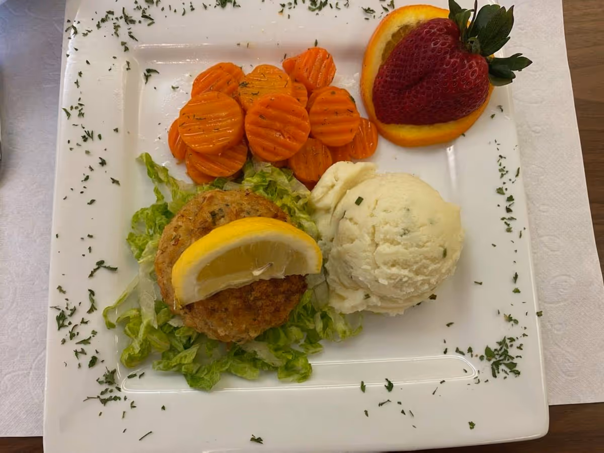 A plated meal consisting of a crab cake topped with a lemon wedge on a bed of shredded lettuce, a scoop of mashed potatoes with herbs, cooked sliced carrots, and a garnish of a strawberry and an orange slice on a square white plate with sprinkled herbs around the edges.