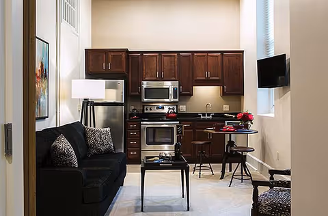 Open-plan living area with a dark sofa and coffee table facing a compact kitchen with dark wood cabinets and stainless steel appliances.