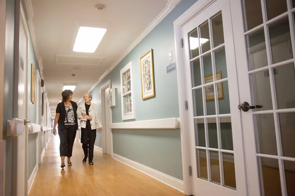 Two women walking and talking down a well-lit hallway with light blue walls, wooden flooring, framed artwork, and white handrails. There are glass-paned double doors on the right side.