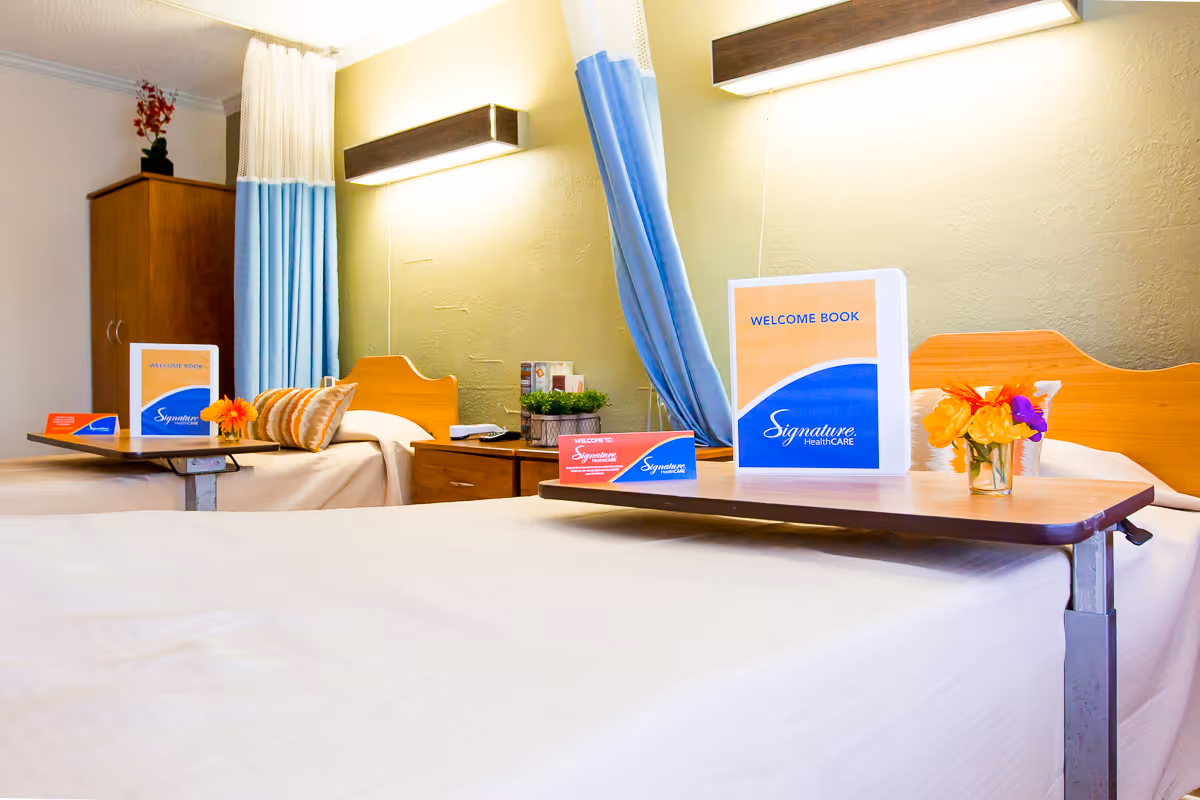 A bright and clean room with two hospital-style beds separated by a blue privacy curtain. Each bed has a wooden headboard and a small overbed table with a welcome book and a small vase of flowers. There is a wooden cabinet in the background and soft lighting above each bed.