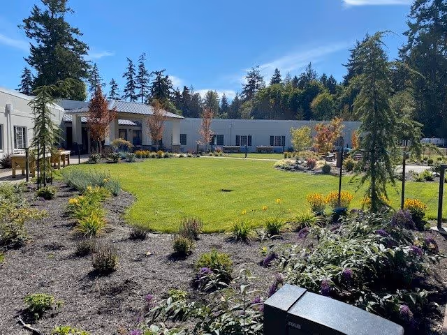 A landscaped outdoor garden area with a circular green lawn surrounded by flower beds and small trees. The garden is enclosed by a single-story white building with multiple windows. The sky is clear and blue, and tall evergreen trees are visible in the background.