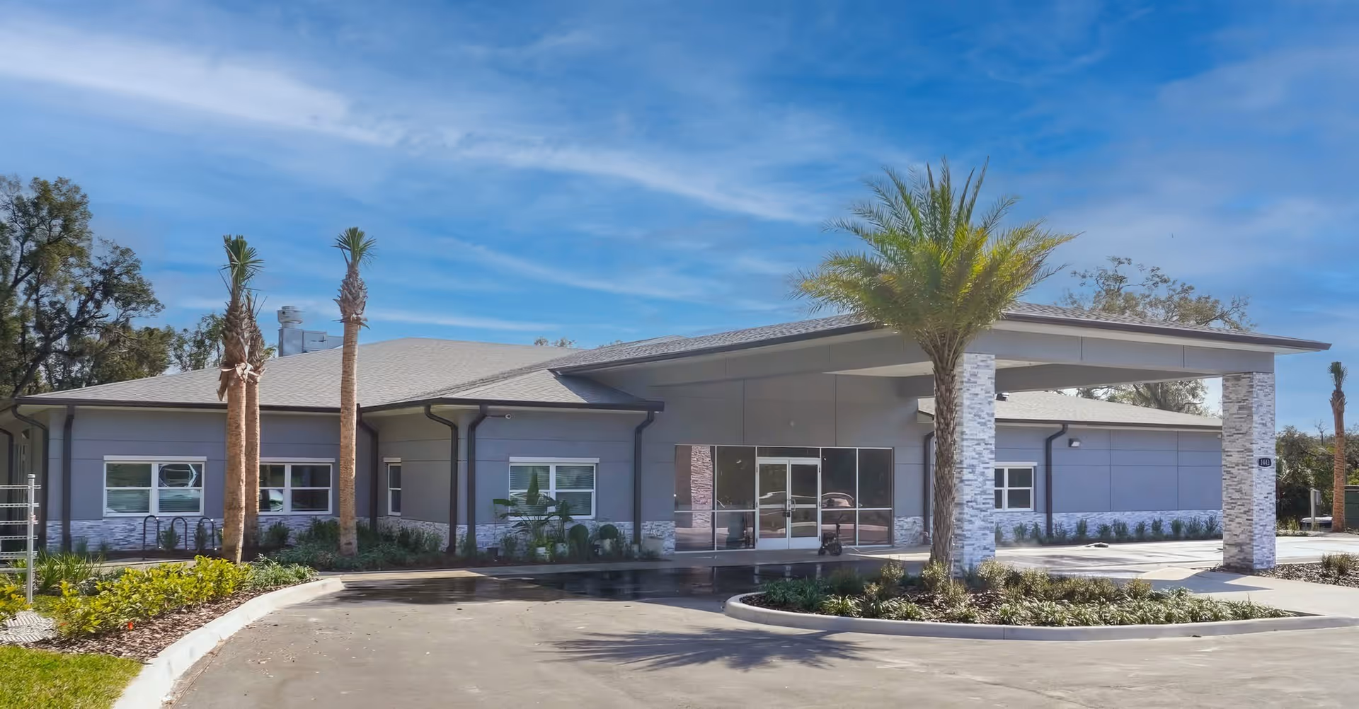 Exterior view of Apopka Life Care Center, a single-story building with a covered entrance supported by stone pillars, surrounded by palm trees and landscaping under a blue sky with some clouds.