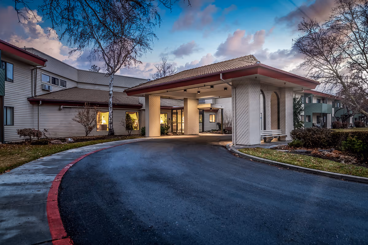 Exterior view of Arbor Village At Hillcrest senior living facility at dusk, showing the main entrance with a covered drop-off area, surrounding buildings, trees without leaves, and a curved driveway with a red curb.