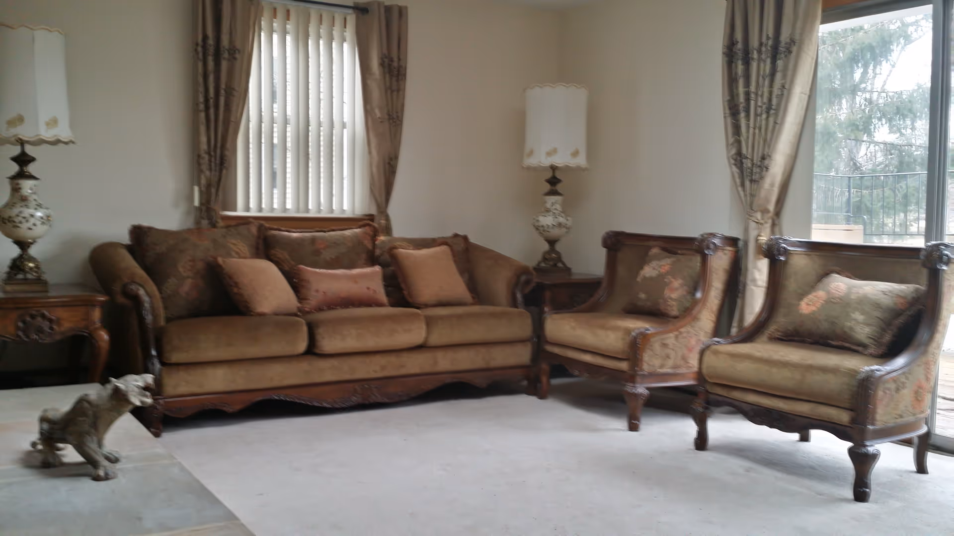 A living room with a brown upholstered sofa and two matching armchairs, each with decorative cushions. There are two ornate table lamps on wooden side tables, beige curtains with floral patterns, a window with vertical blinds, and a glass door leading outside. A small dog statue is placed on the floor near the sofa.