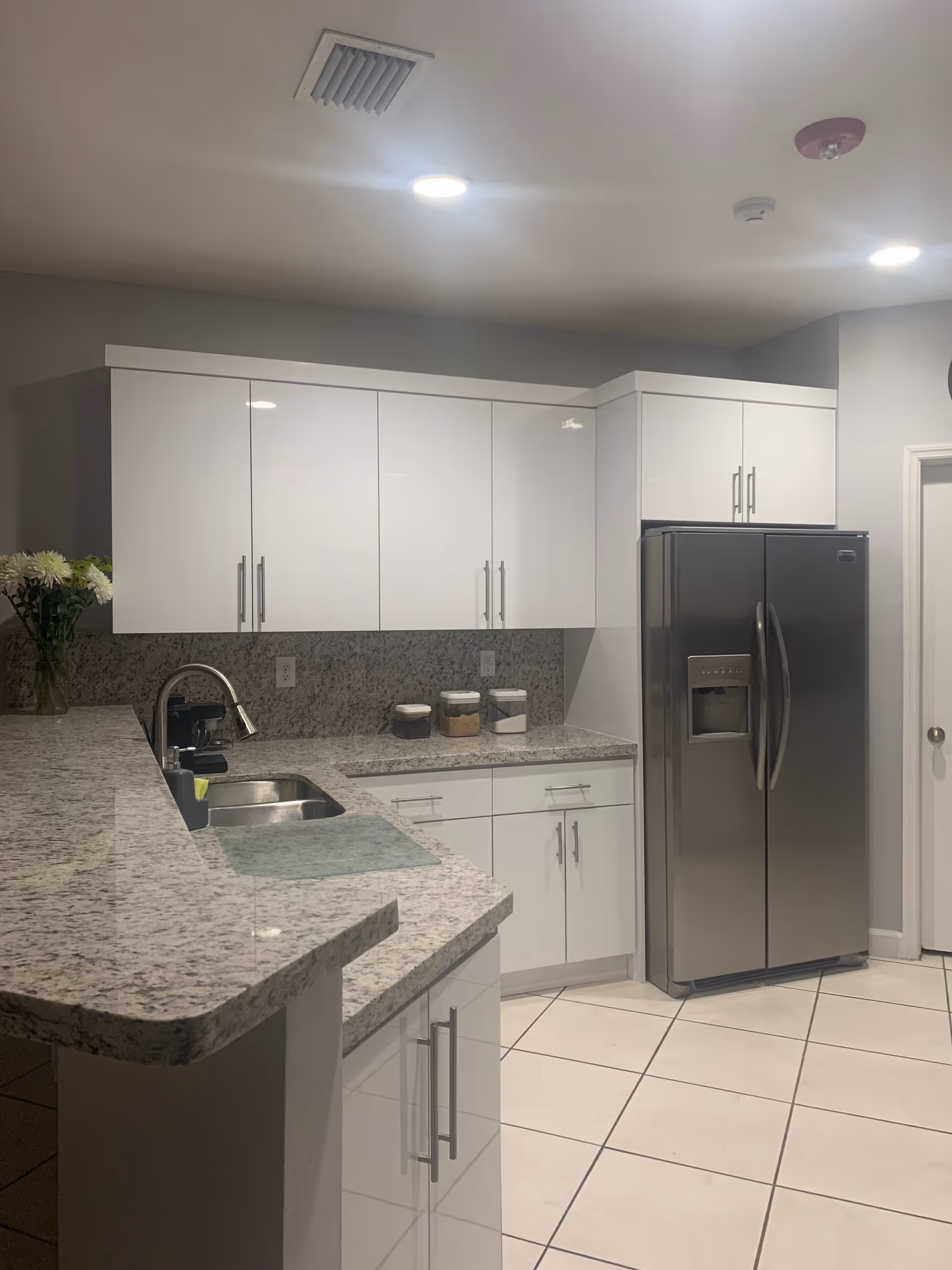 Modern kitchen with white cabinets, granite countertops, a stainless steel refrigerator, and a double sink. There are three jars on the counter and a vase with flowers in the corner.