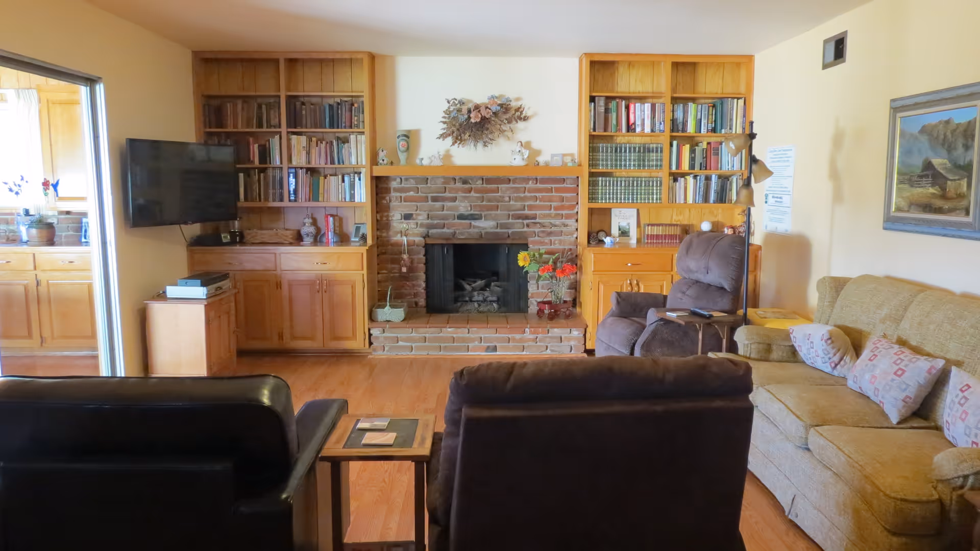 A cozy living room with a brick fireplace in the center flanked by wooden built-in bookshelves filled with books and decorative items. There is a flat-screen TV mounted on the left wall near an open doorway leading to a kitchen. The room has a brown recliner, a beige sofa with patterned pillows, and a black leather chair. A small wooden side table is placed between the chairs, and a floor lamp stands next to the recliner. A framed painting hangs on the right wall.