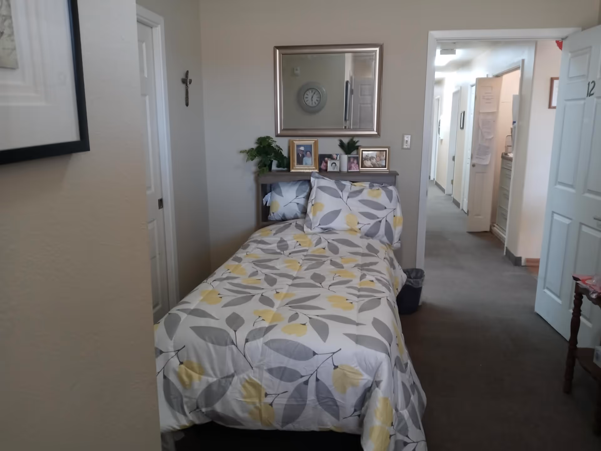 Small bedroom with a single bed dressed in gray and yellow leaf-patterned bedding, a mirror and framed photos above the headboard, and a hallway visible through an open door.